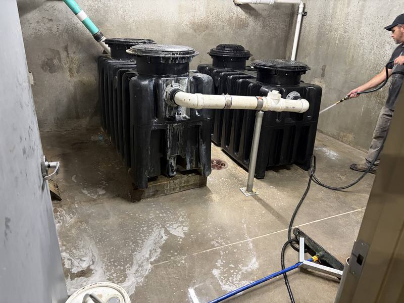 TCS tech pressure-washing a utility room with four black grease interceptors and white PVC piping