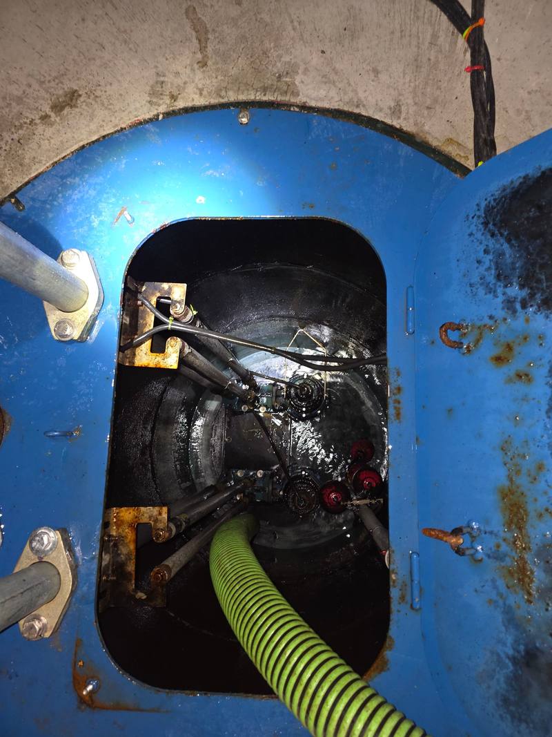 Top-down view into an open blue steel grease tank hatch with a green suction hose going in